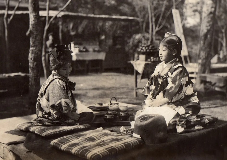 Two girls in kimono enjoying tea outdoors in early 20th-century Japan.