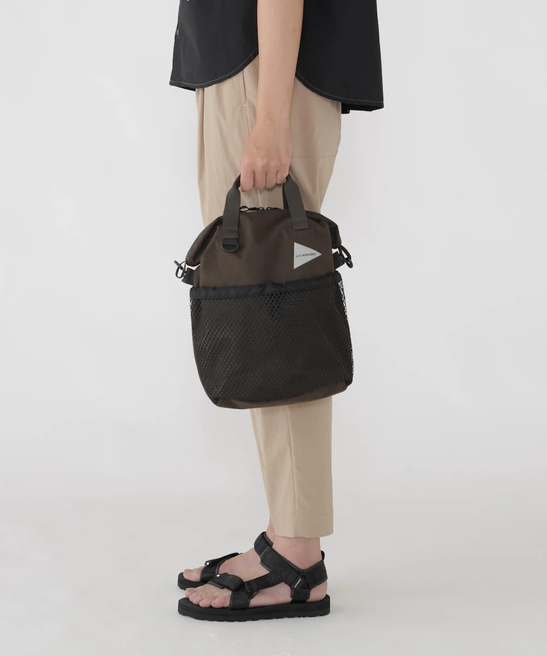 Sac utilitaire and wander en nylon avec poche filet extérieure, porté à la main avec sandales techniques, illustrant l’accessoire fonctionnel emblématique du style gorpcore japonais.