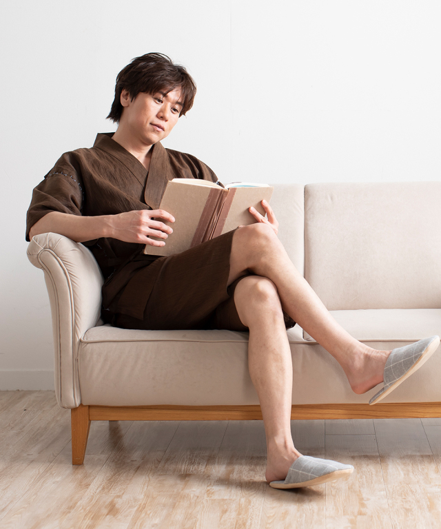 Homme portant un jinbei marron, assis sur un canap&eacute; et lisant un livre &agrave; la maison.