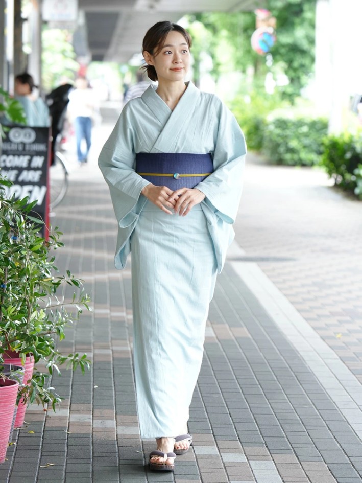 Femme portant un yukata bleu clair crois&eacute; gauche sur droite, maintenu par un obi bleu marine nou&eacute; &agrave; la taille, avec des geta aux pieds.