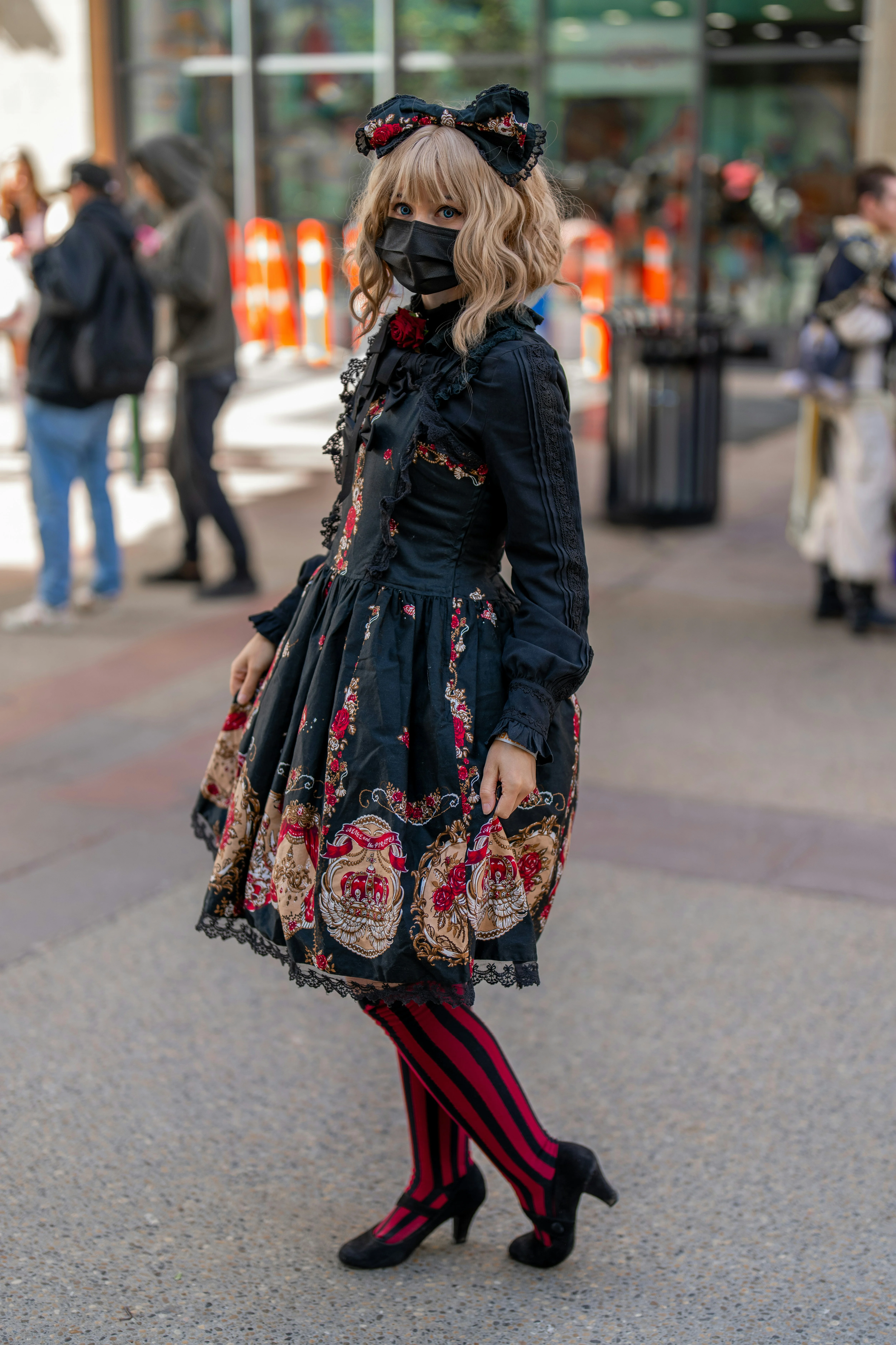 Persona posando con un vestido de estilo Lolita g&oacute;tico en colores negro y rojo, con detalles florales y encajes. Completa el look con una diadema a juego, medias a rayas y zapatos de tac&oacute;n, en un entorno urbano.
