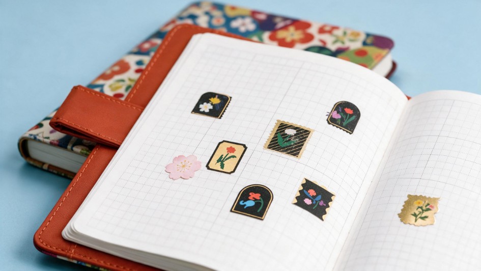 A close-up flat lay showing an open Hobonichi planner with gridded paper decorated with small floral stickers. The open book rests on a rust-orange cover with a pen loop and a colorful floral patterned cover, highlighting the variety of different Hobonichi planner formats and customization options available.