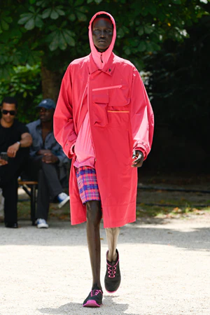 Silhouette White Mountaineering en manteau technique rouge vif à capuche, porté sur short à carreaux et sneakers outdoor, illustrant l’approche audacieuse et stylisée du gorpcore japonais.